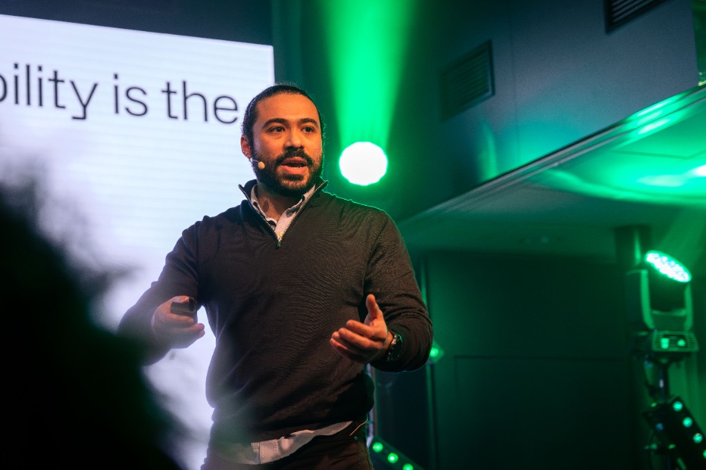 Aly Metwaly delivering the opening keynote at Verge at Epicenter, on stage with green lighting and a headset microphone